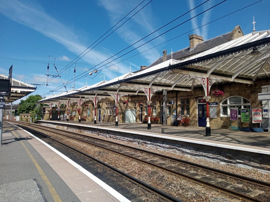 Bus station in Skipton, Yorkshire, featuring a stone building, platforms, and decorative canopies under a blue sky.