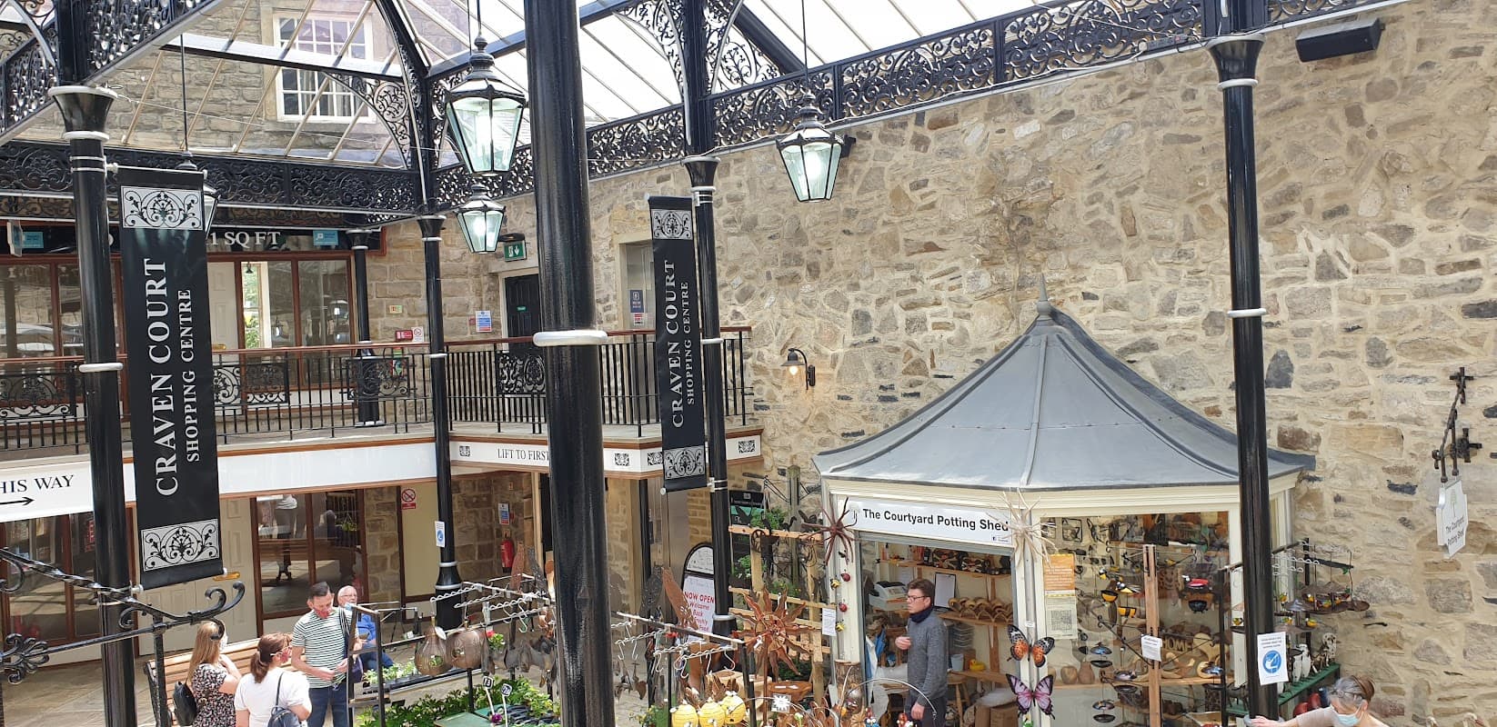 Indoor market featuring stone walls, decorative ironwork, stalls with crafts, and shoppers browsing.