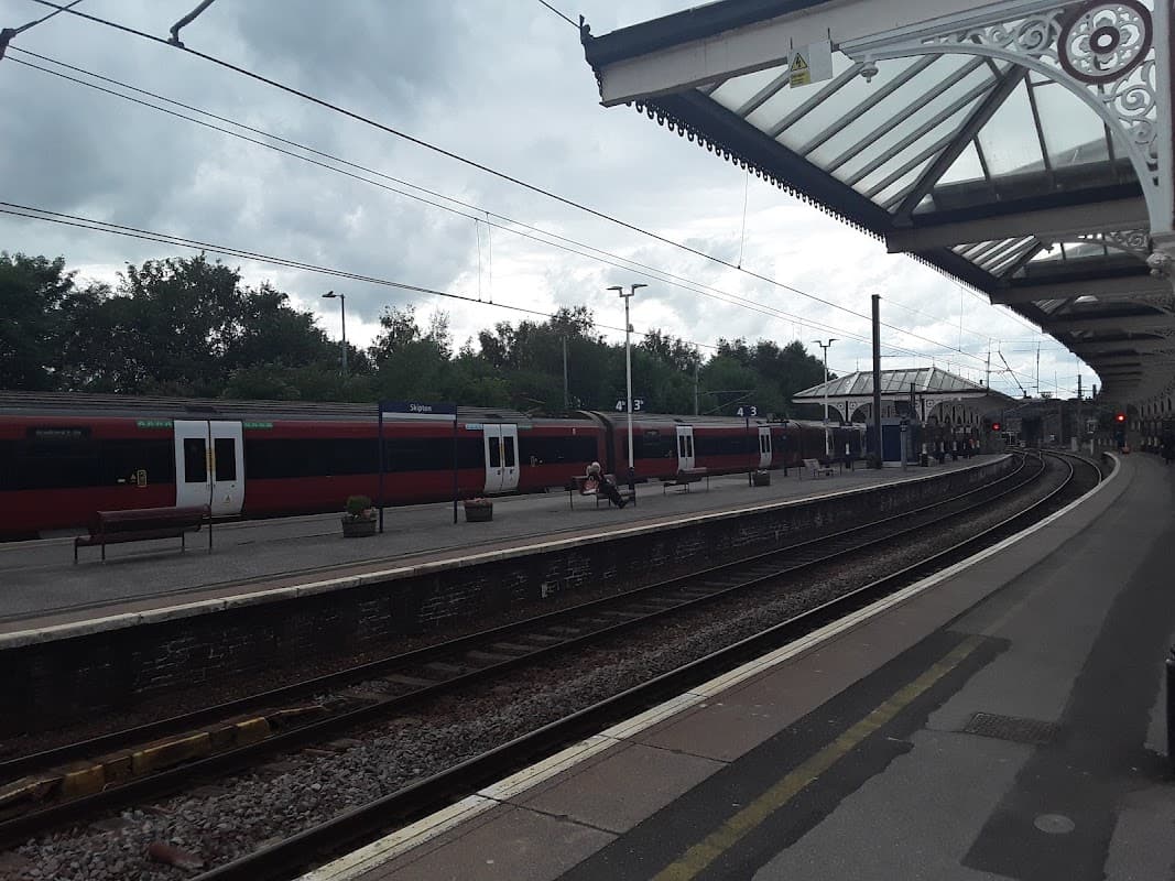 Skipton Railway Station features two red trains, a covered platform, and a cloudy sky in Yorkshire.