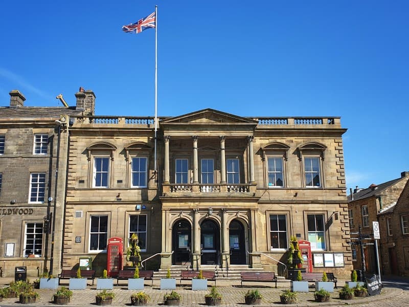 Skipton Town Hall features classical architecture, a Union Jack flag, and flower planters in a sunny square.