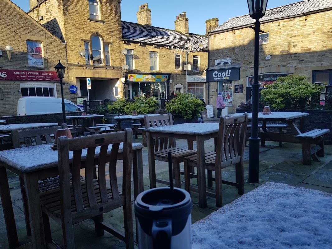 Outdoor seating area with wooden tables and chairs, a coffee cup in the foreground, and stone buildings in the background.