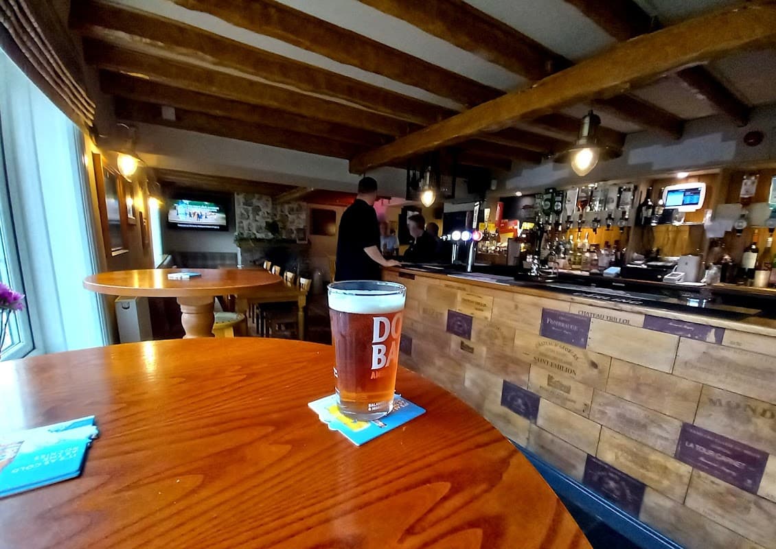A pint of amber beer sits on a wooden table, with a rustic bar and bartender in the background.