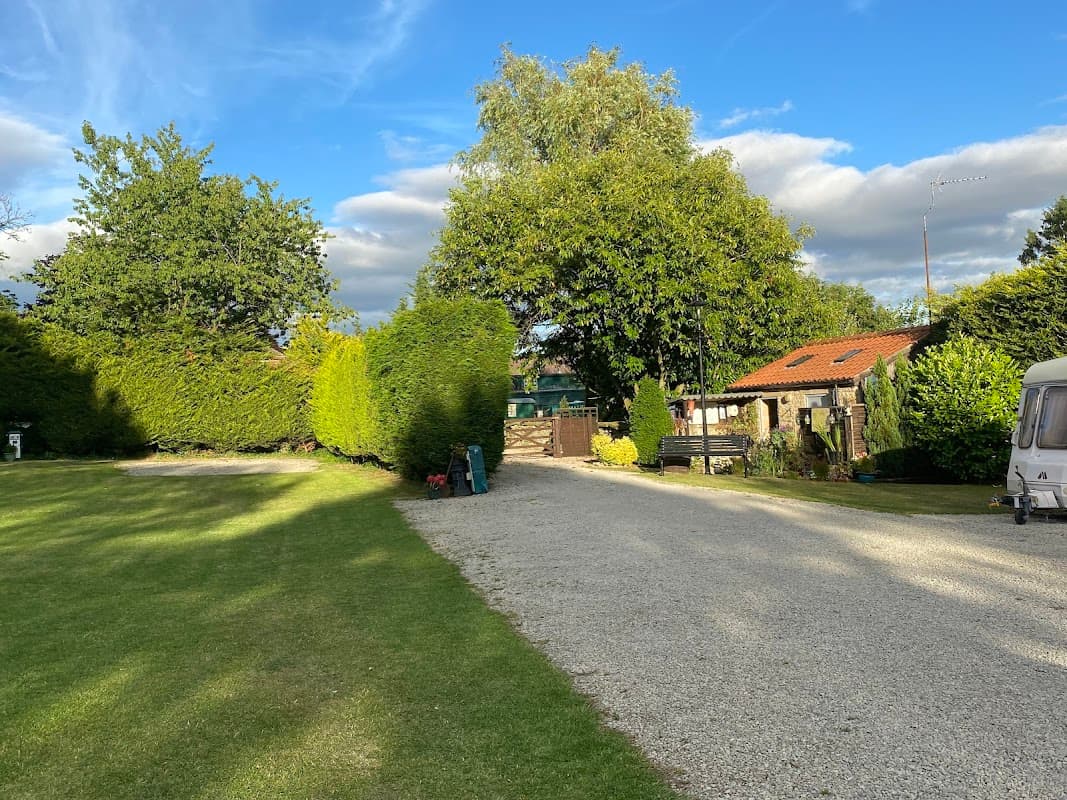 Gravel pathway leading through lush greenery and trees, with a cozy building and blue sky in the background.