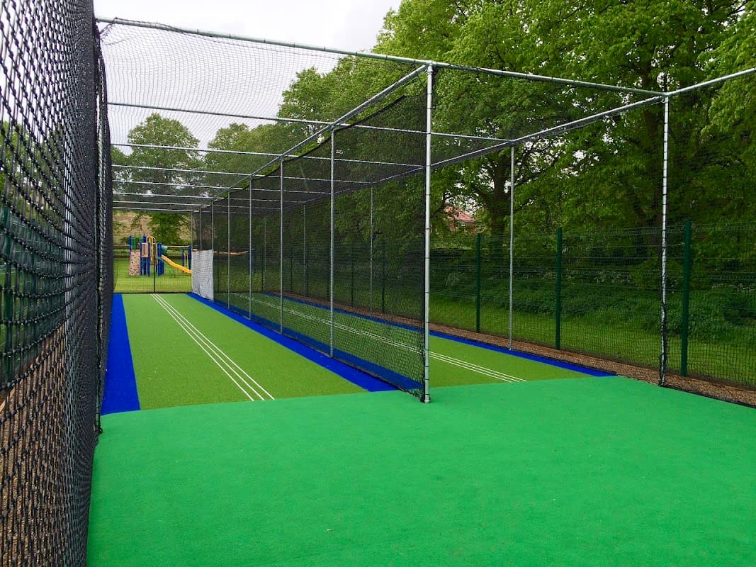 Cricket practice nets with green turf, surrounded by trees and a playground in the background.