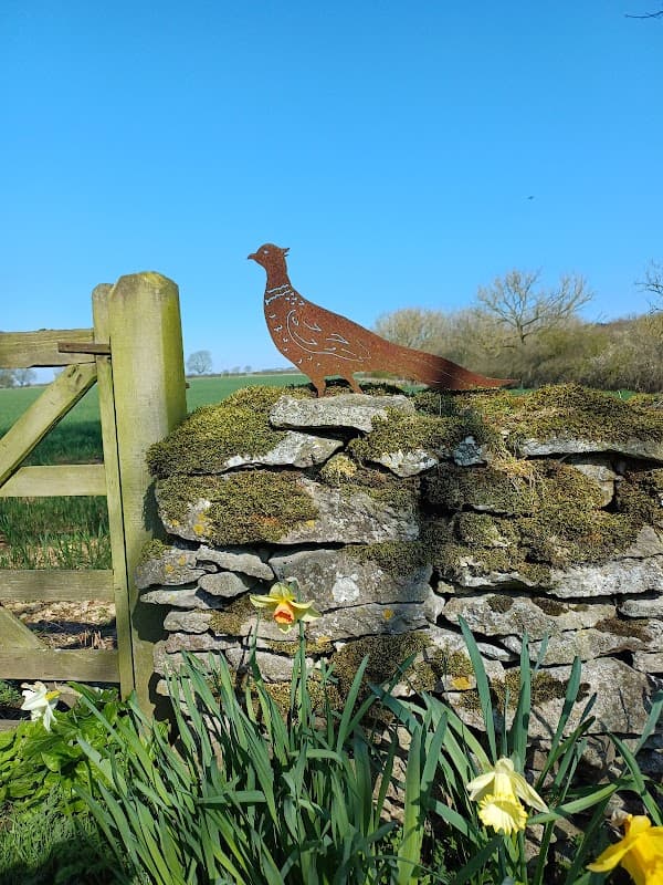 A metal pheasant sculpture on a stone wall, surrounded by daffodils and a clear blue sky in Snainton, Yorkshire.
