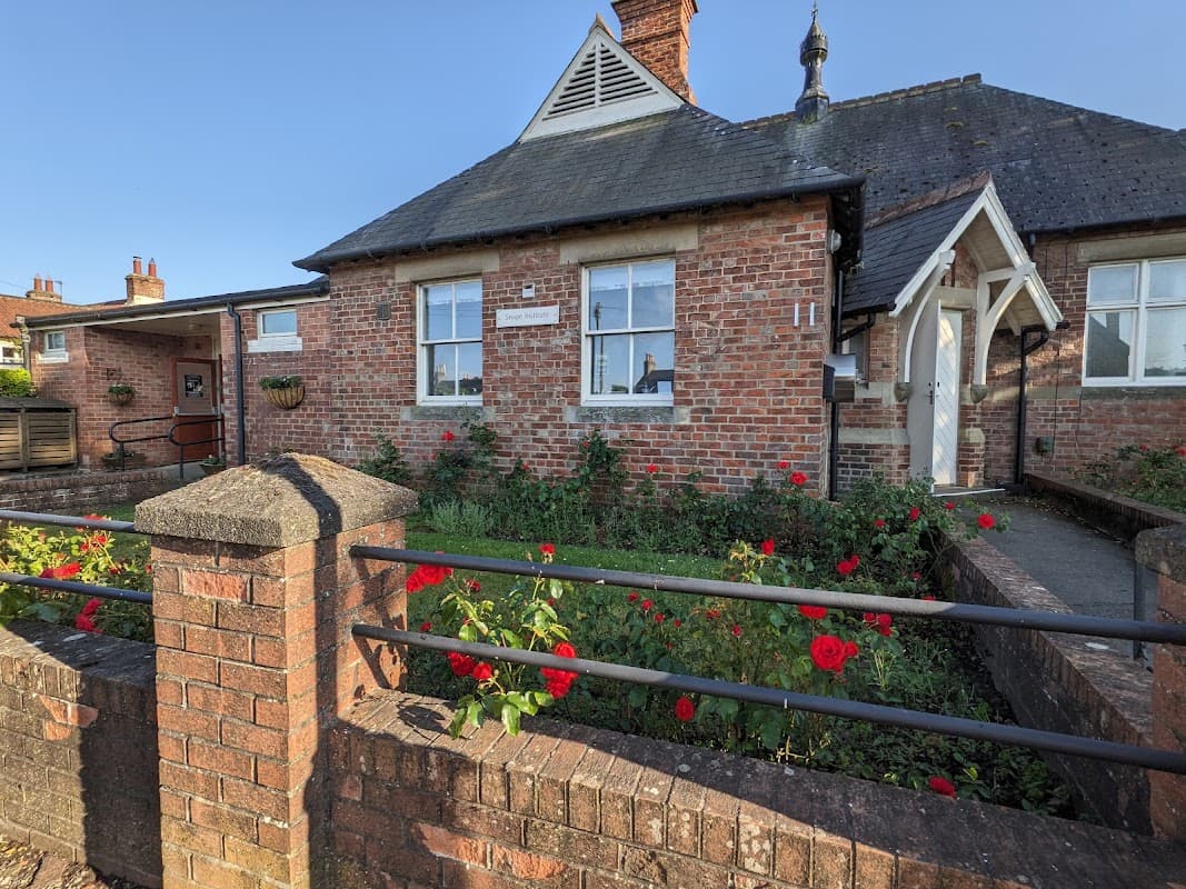 Brick building with a gabled roof, white trim, and a garden of red roses in front. Clear blue sky above.