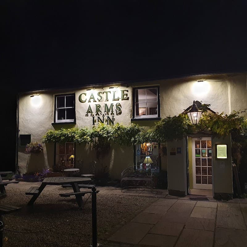 The Castle Arms Inn, a cozy bar with illuminated signage, outdoor seating, and lush greenery at night.