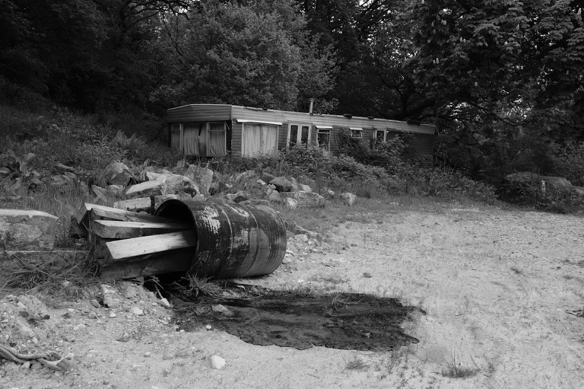 A derelict caravan surrounded by trees, with a rusty barrel and wooden planks in the foreground on a gravel path.