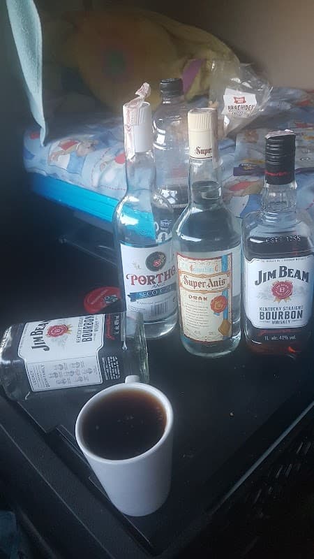 Bottles of various spirits and a cup of dark liquid on a table, with a colorful bedspread in the background.