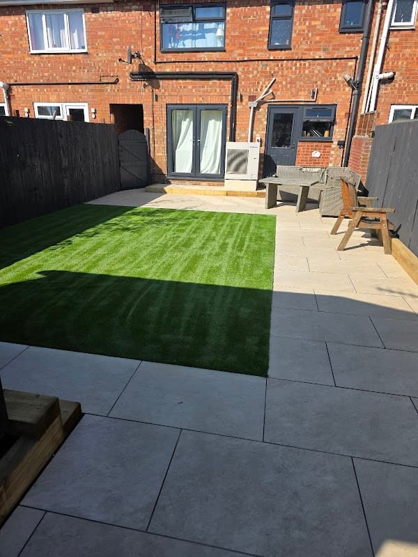 Lush green artificial grass area with tiled patio, wooden chairs, and a wooden fence in a sunny garden setting.
