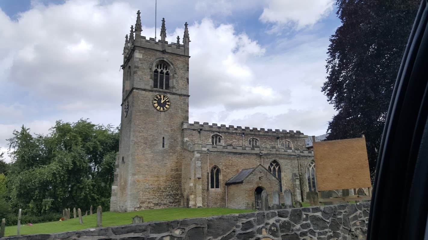 Historic church with a tall clock tower, surrounded by trees and a stone wall, under a partly cloudy sky.