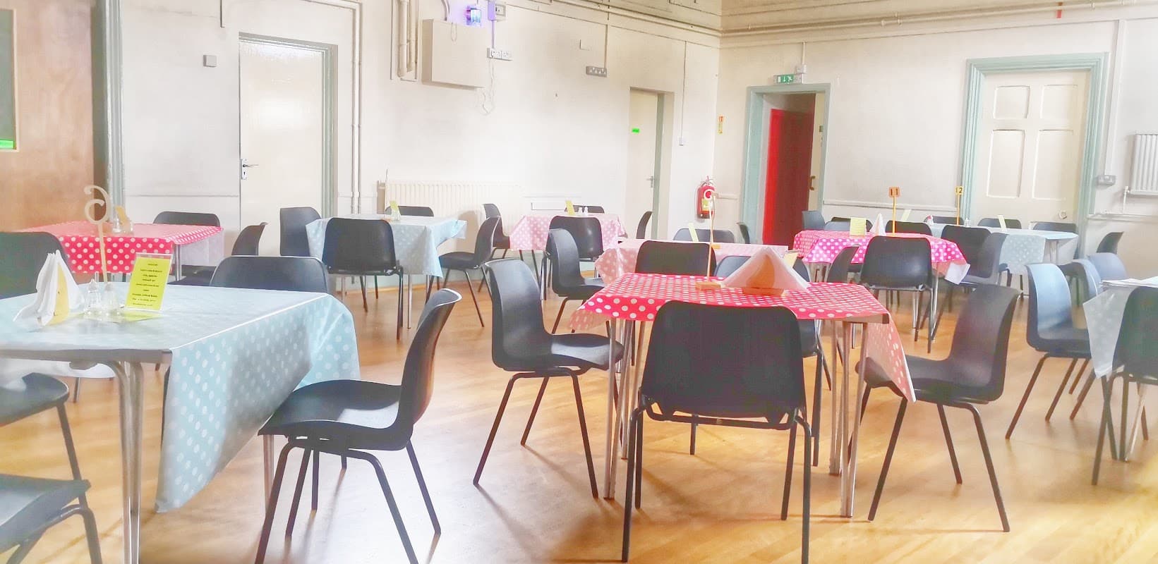 Brightly lit hall with tables set in blue and red checkered cloths, surrounded by black chairs and wooden flooring.