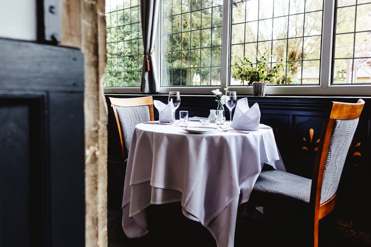 Elegant dining table set with white tablecloth, glassware, and napkins, surrounded by large windows and greenery.