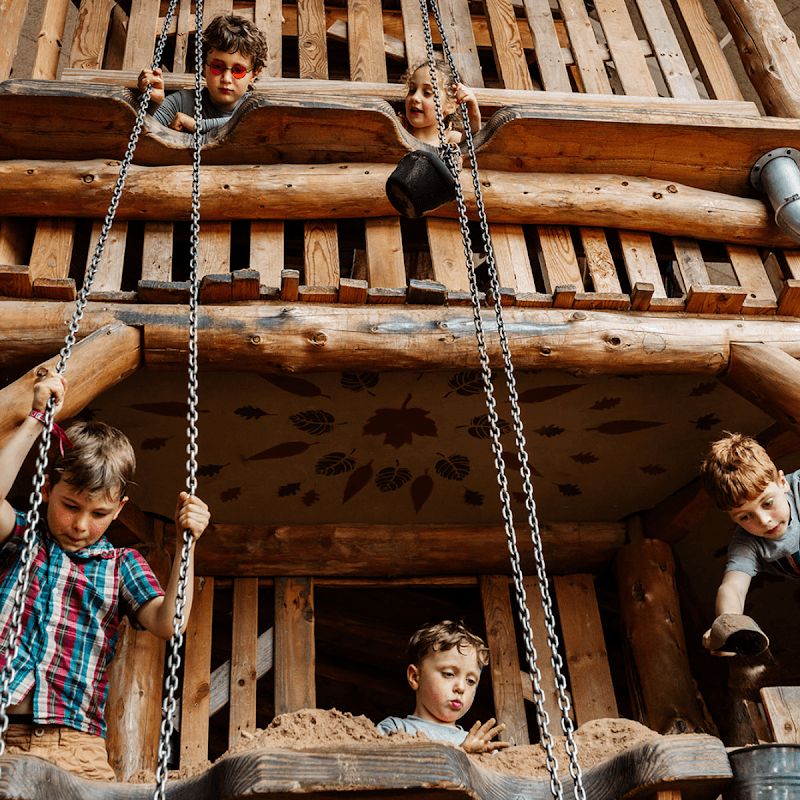 Children play on swings and a wooden structure, with sand below, in a rustic outdoor area at William's Den.
