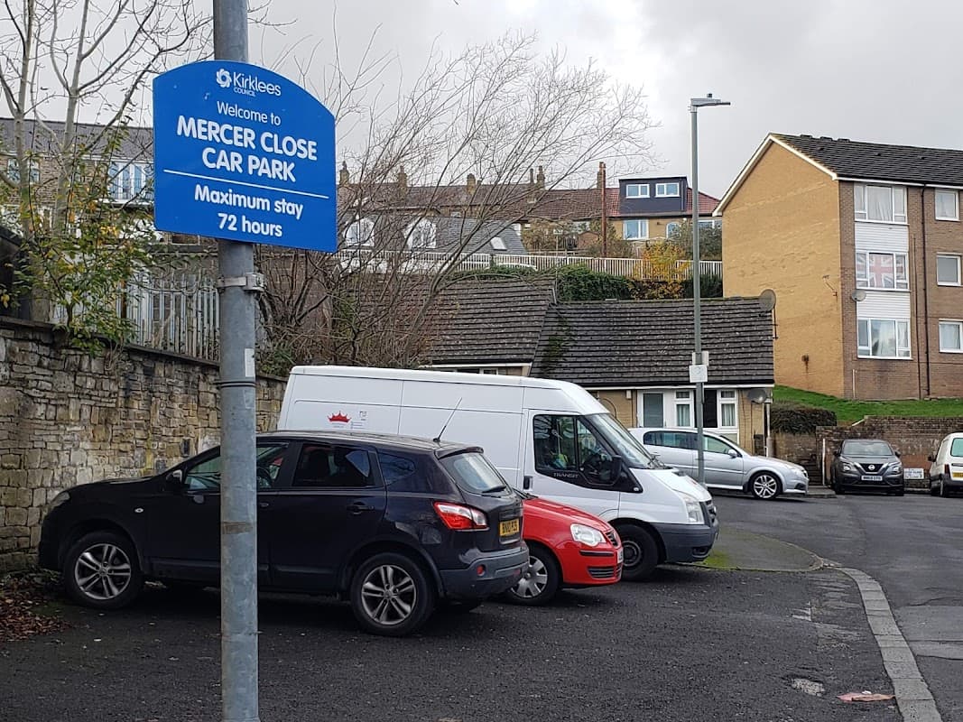 Sign for Mercer Close Car Park with parked cars and residential buildings in the background on a cloudy day.