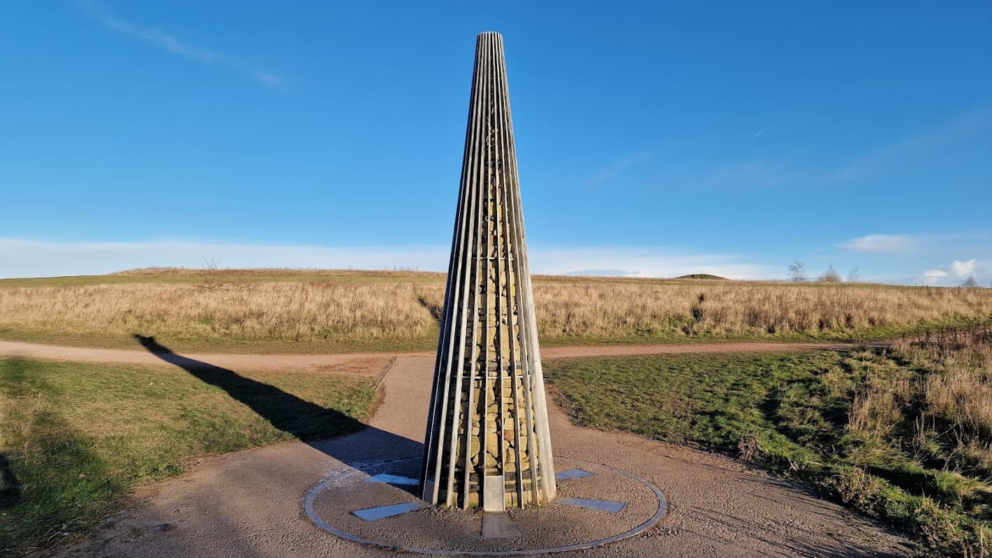 A tall, pointed wooden structure stands on a path, surrounded by grassy fields under a clear blue sky.