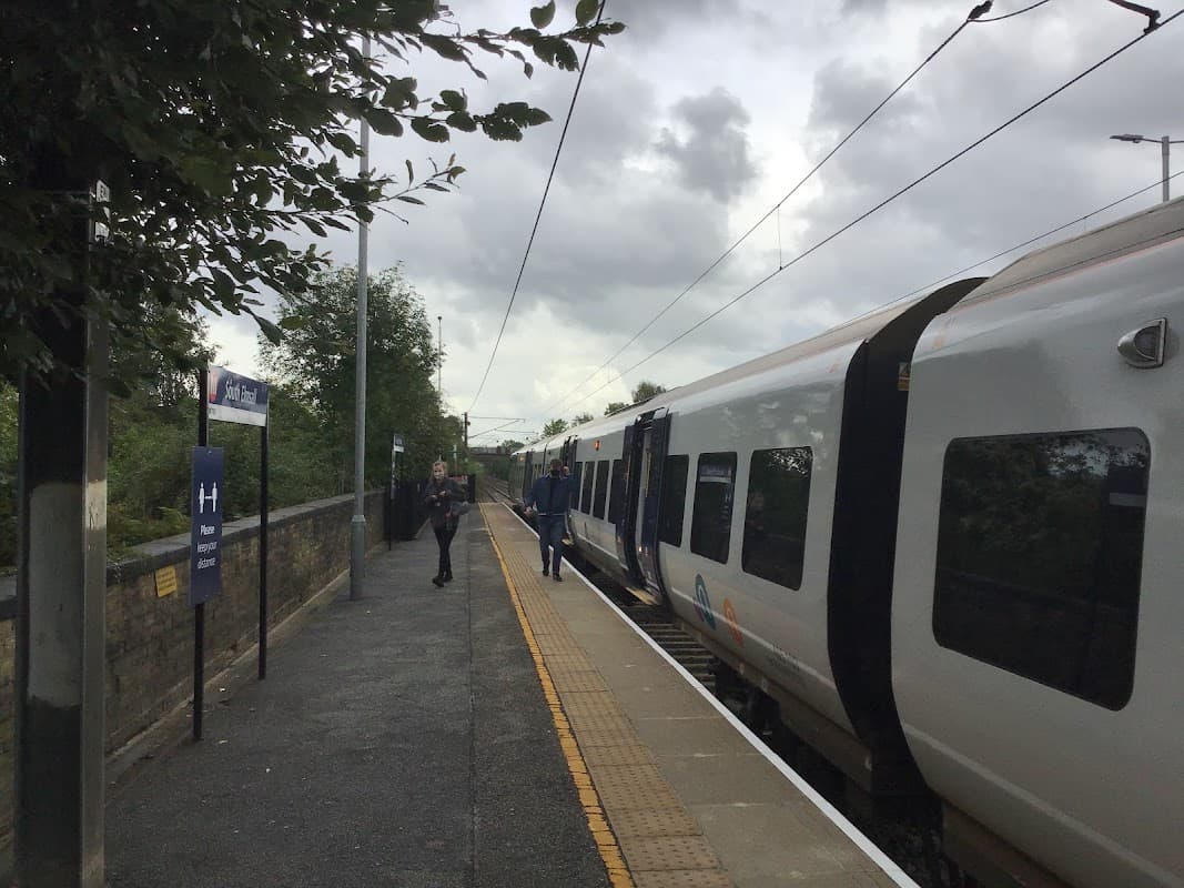 Bus stop at South Elmsall with passengers waiting and a train arriving, under a cloudy sky.