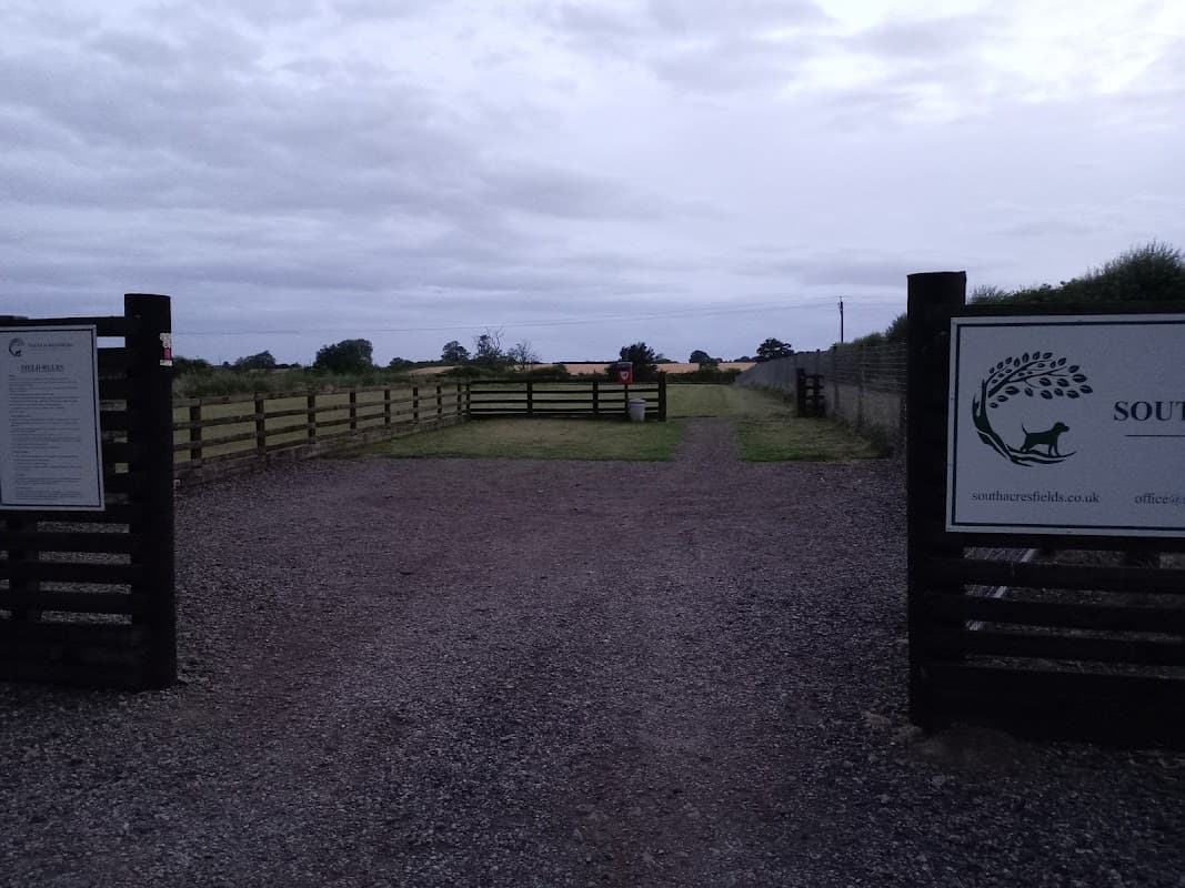 Entrance to South Acres Dog Fields with wooden gates, gravel path, and open grassy fields under a cloudy sky.