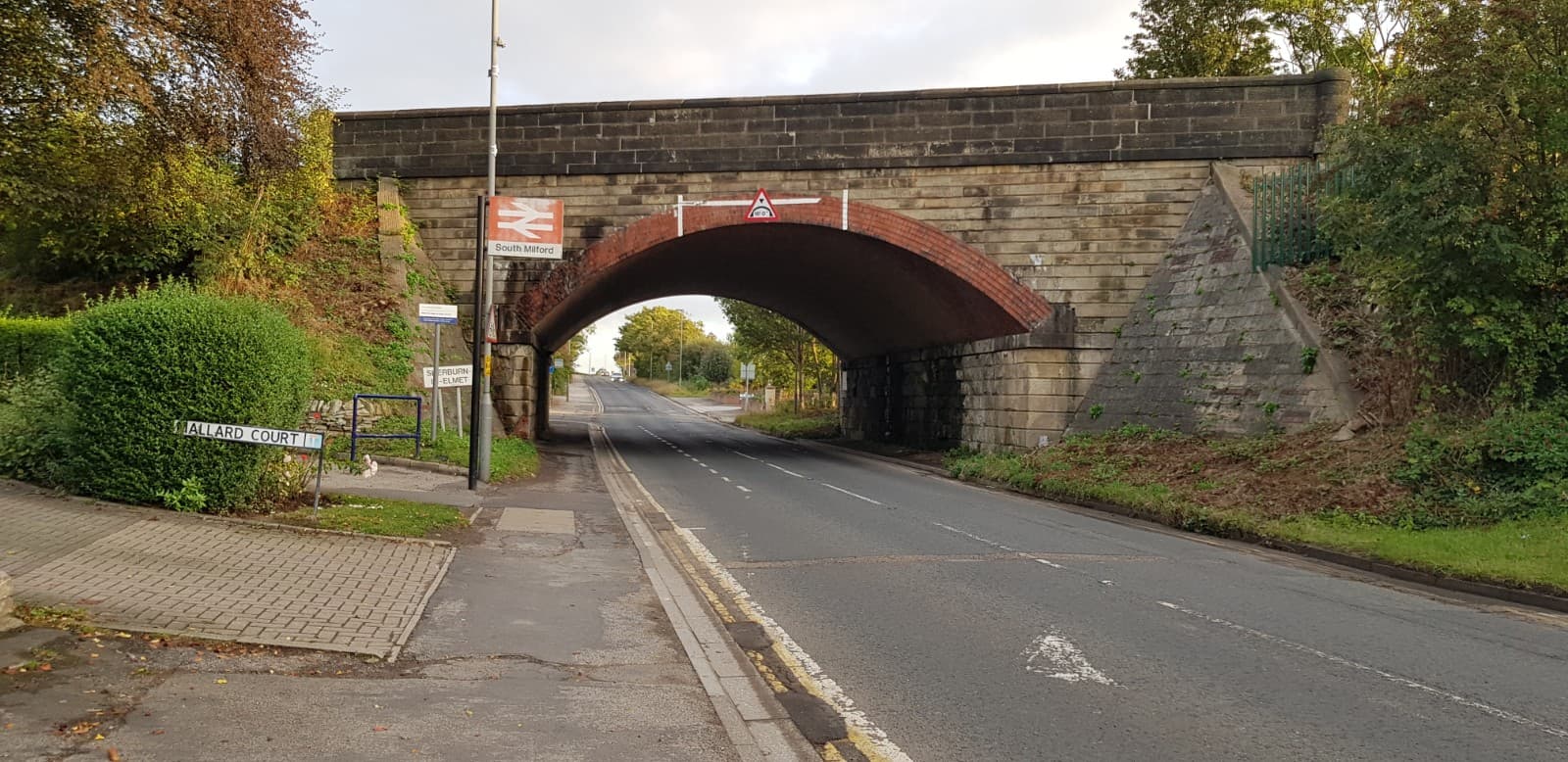 Bus stop near a brick railway bridge, with trees lining the road and a sign for "Halliard Court" visible.