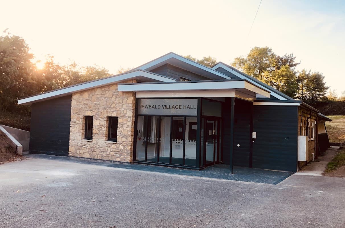 Modern village hall with stone and wooden exterior, large glass entrance, surrounded by greenery and trees.