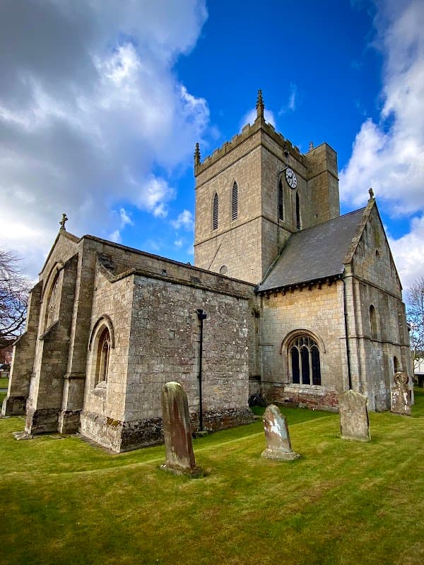 Historic church with a tall clock tower, surrounded by gravestones and lush green grass under a blue sky.