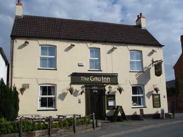 The Gnu Inn pub features a cream-colored exterior, wooden signage, and outdoor seating with flower baskets.