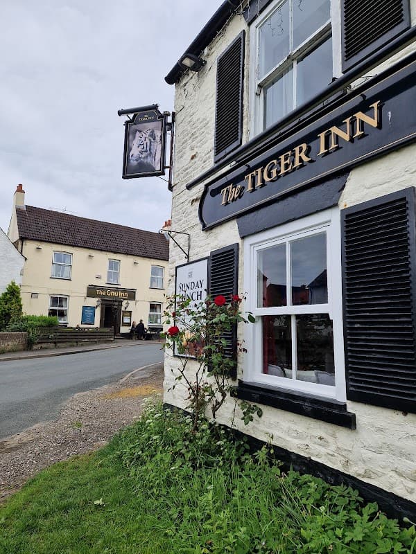 The Tiger Inn sign hangs beside a white building with black shutters, roses in front, and a view of a nearby pub.