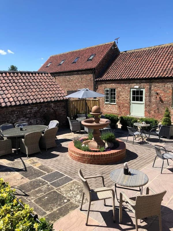 Outdoor seating area with a fountain, surrounded by brick walls and buildings under a clear blue sky.