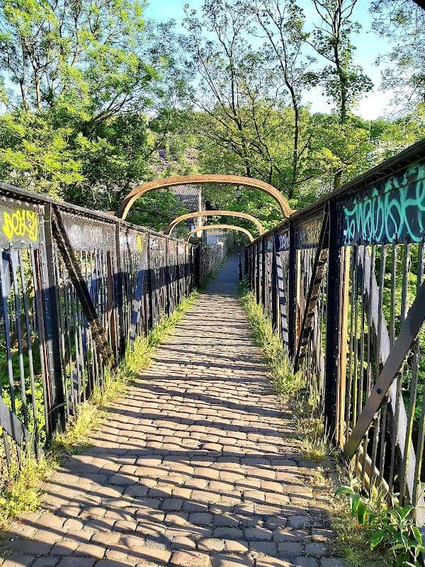 Footpath leading through a bridge surrounded by greenery, with graffiti on the railings and sunlight filtering through trees.