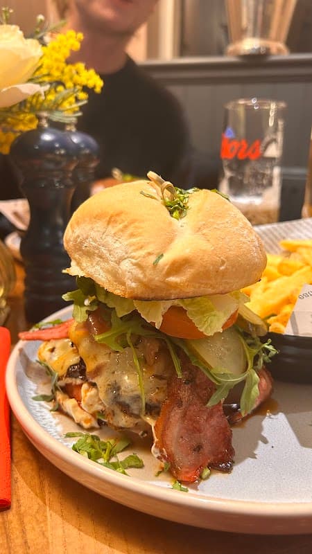 A gourmet burger stacked with bacon, cheese, lettuce, and tomato, served with fries and a flower centerpiece.