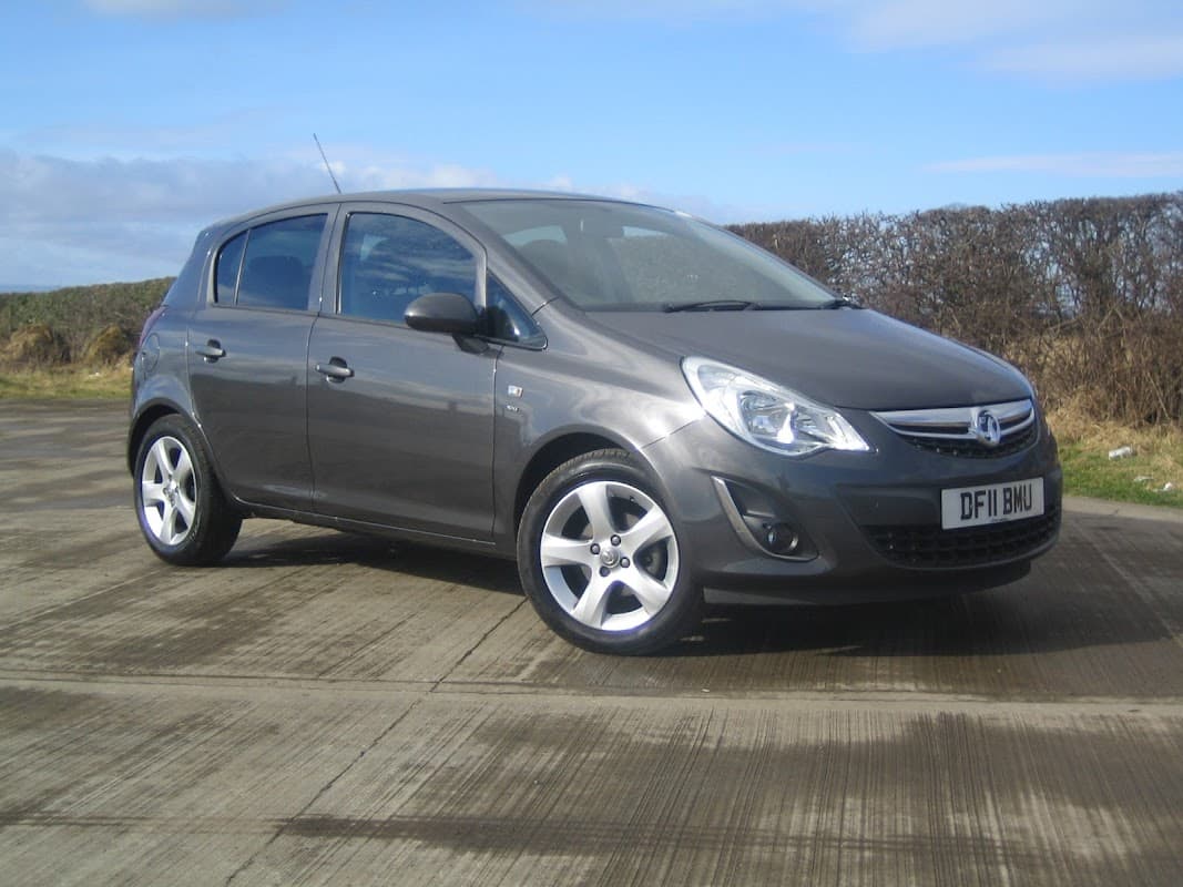 Gray hatchback parked on a concrete surface, with a blue sky and hedgerow in the background.