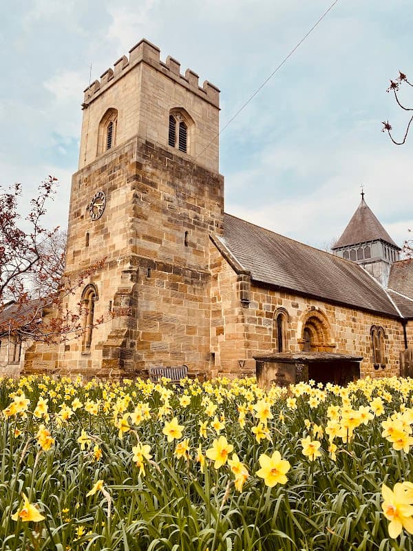 Sowerby Parochial Hall surrounded by blooming daffodils under a cloudy sky, featuring a stone tower and arched windows.