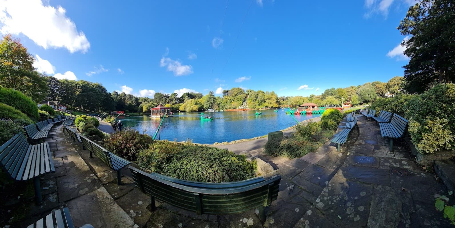 Lush greenery surrounds a tranquil lake with colorful boats, benches lining the path under a bright blue sky.