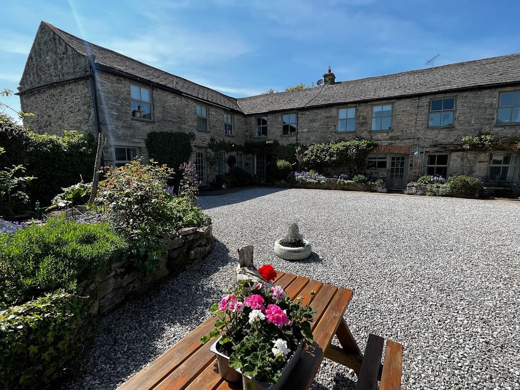 Charming stone cottages with a gravel courtyard, surrounded by greenery and flowers under a clear blue sky.