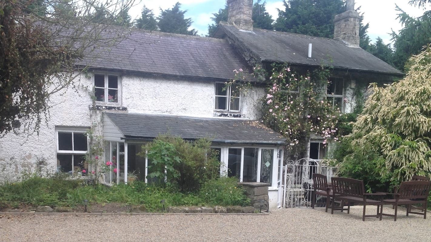 Charming white cottage with climbing roses, surrounded by greenery and outdoor seating in a tranquil setting.