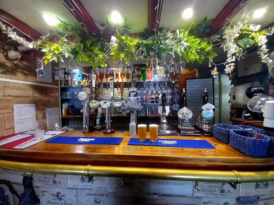 Bar counter with taps, glasses, and two pints; greenery decor above in a cozy pub setting.