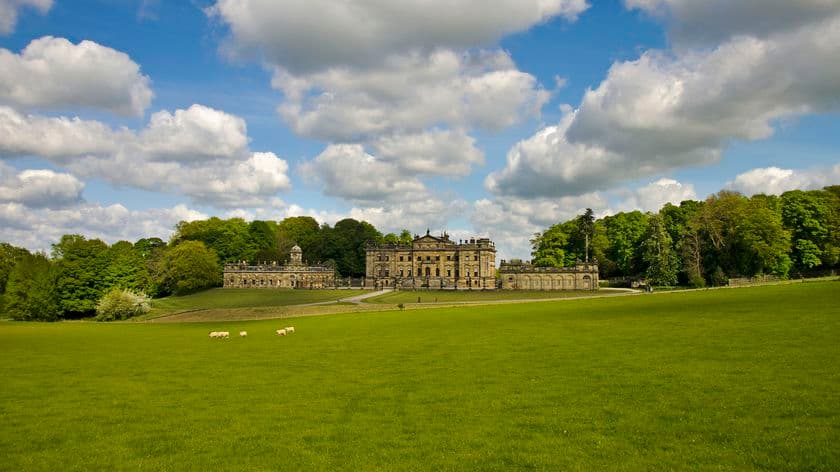 Lush green fields with grazing sheep, a grand historical building, and a backdrop of trees under a partly cloudy sky.