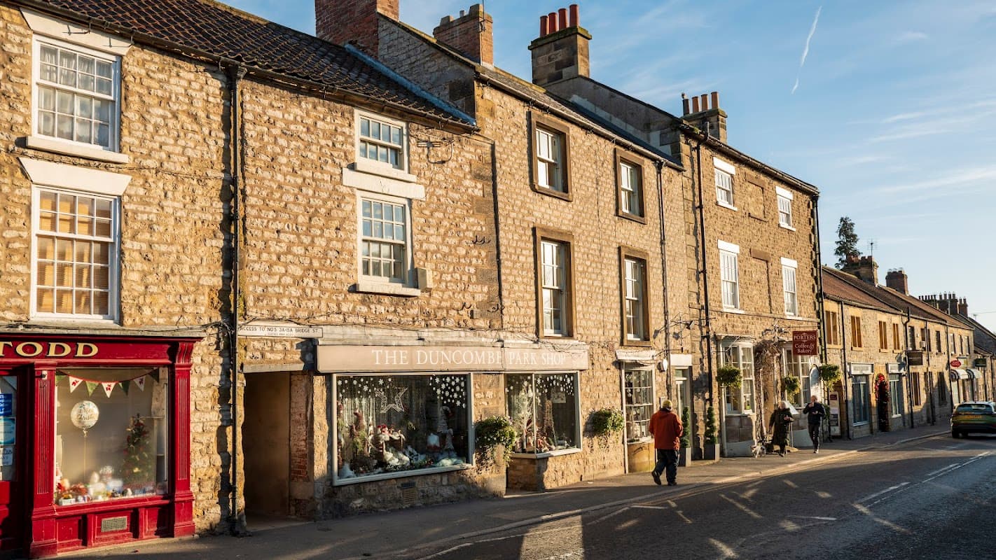 Historic stone buildings line a street, featuring the Duncombe Park Shop with festive decorations and shoppers outside.
