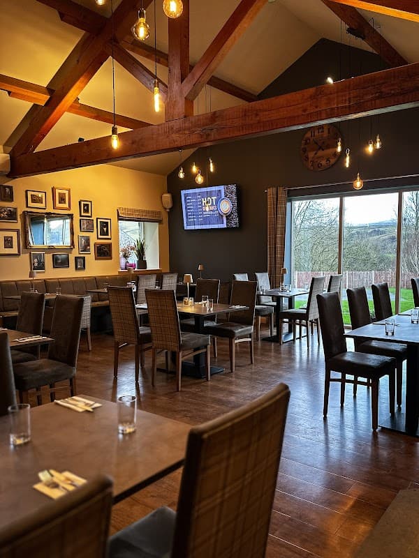 Rustic dining area with wooden beams, stylish chairs, and large windows overlooking greenery. Wall adorned with framed photos.