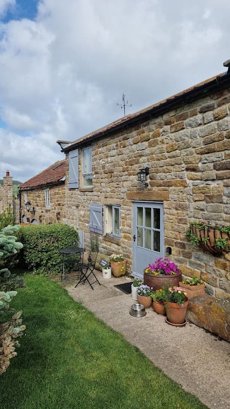 Charming stone cottage with blue shutters, potted flowers, and a small outdoor seating area on a grassy hillside.