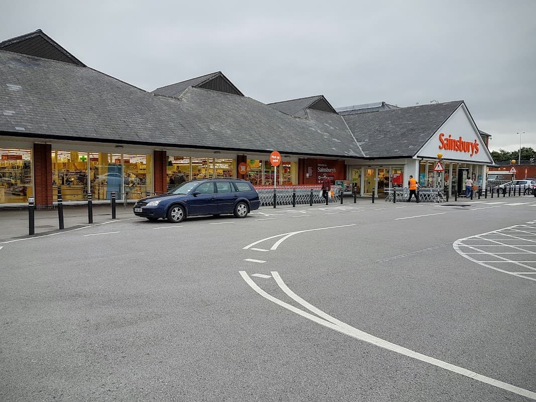 Sainsbury's Petrol Station with a gray car parked outside, surrounded by a spacious parking lot and a cloudy sky.