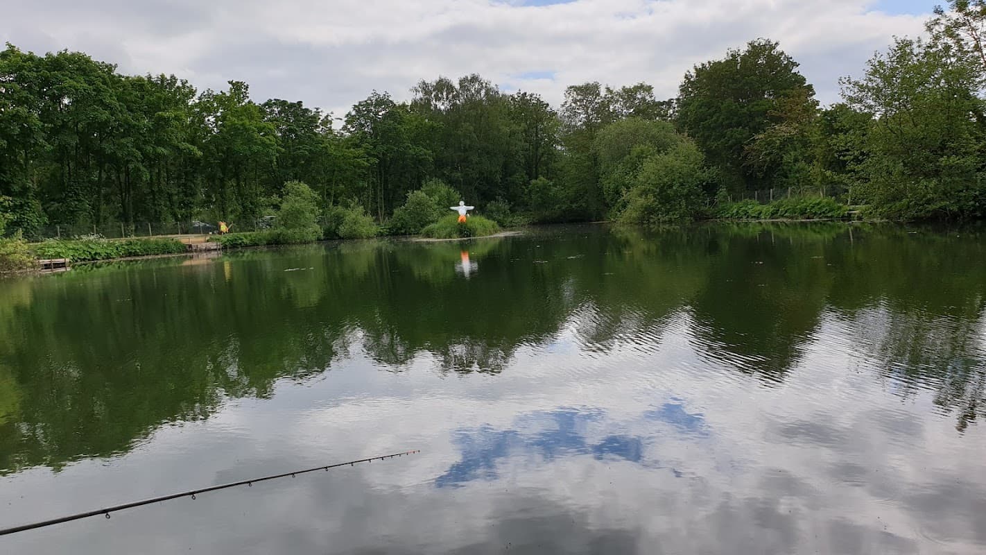 Calm lake reflecting trees and clouds, with a fishing rod in the foreground and a colorful figure on the shore.