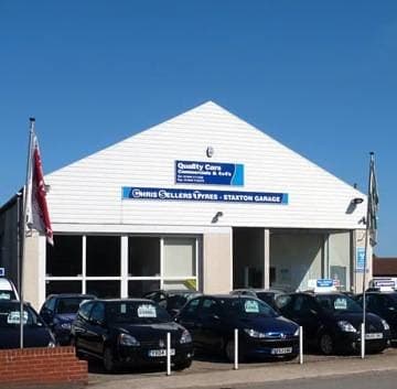 MOT and Used Quality Cars building with multiple parked cars and flags in Staxton, Yorkshire under a clear blue sky.
