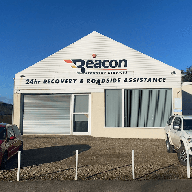 White building with "Beacon Recovery Services" sign, large windows, and gravel parking area. Clear blue sky above.