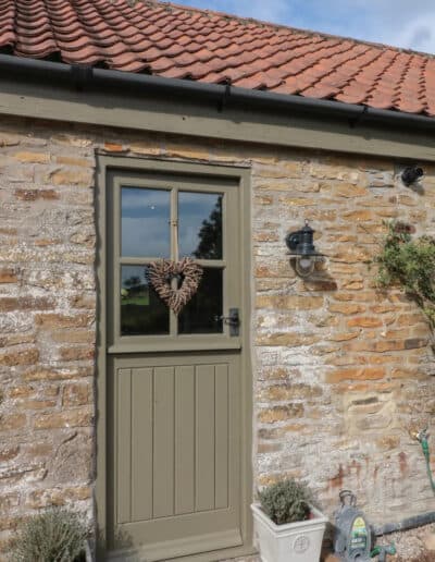 A rustic stone barn with a green door featuring a heart-shaped wreath and a light fixture beside it.