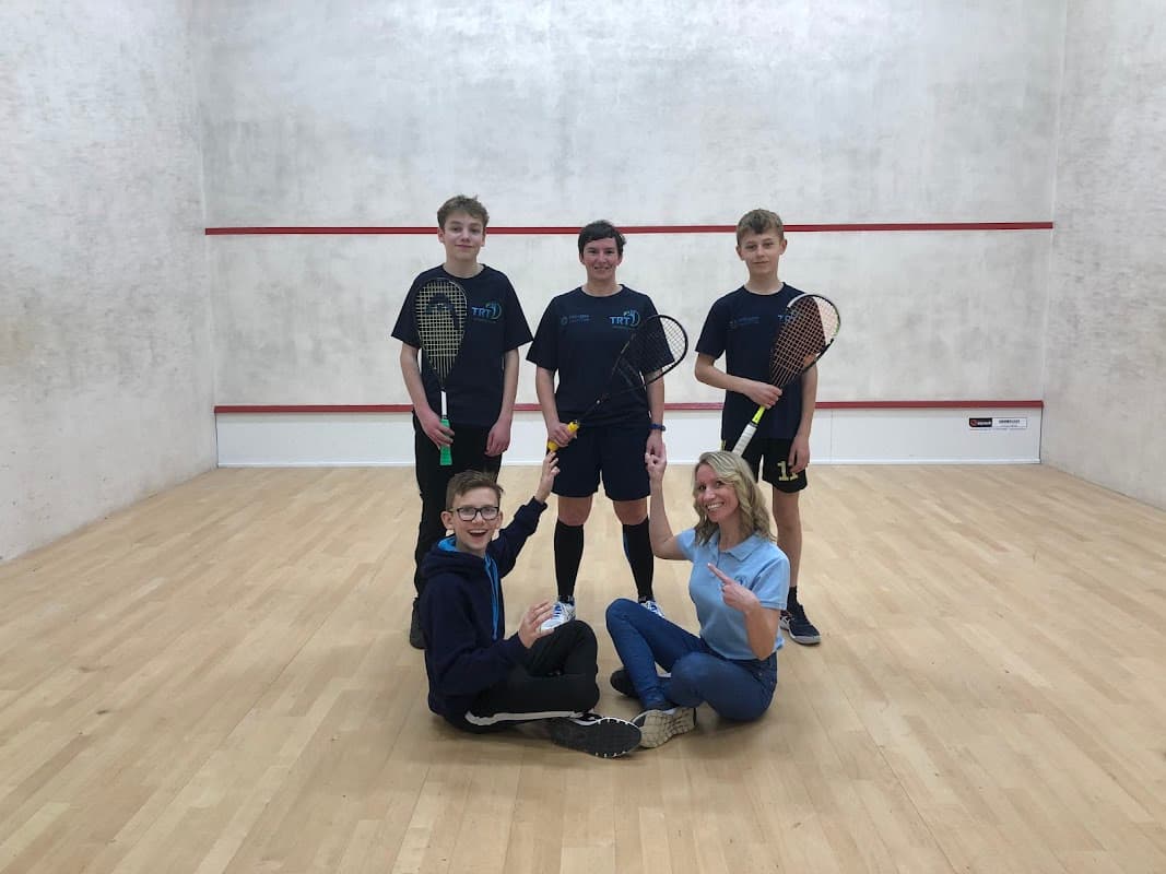 Four young squash players and a coach pose on a squash court, holding racquets and smiling at the camera.
