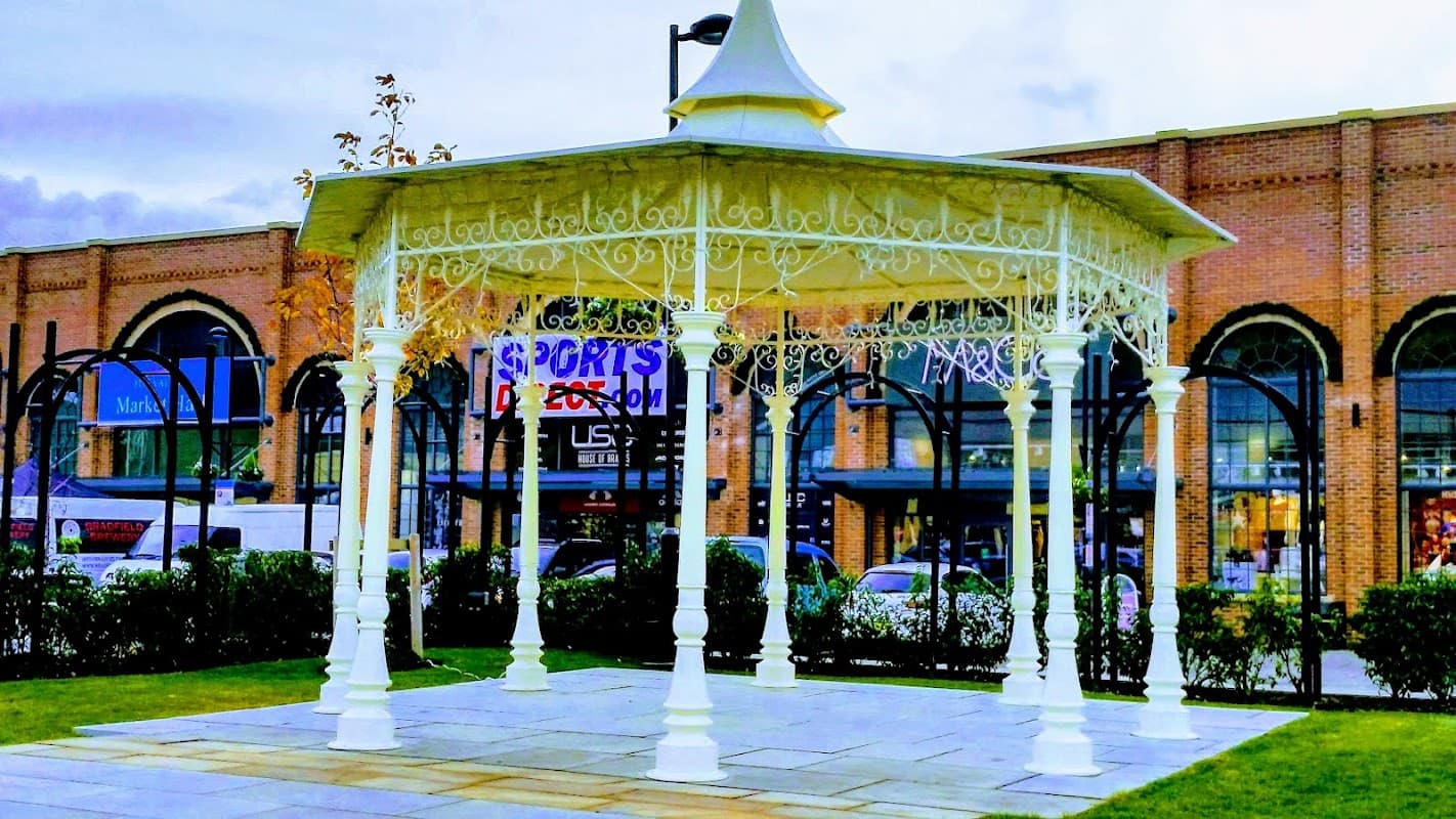 White gazebo in a landscaped area with a view of a "Sports Direct" store in the background.