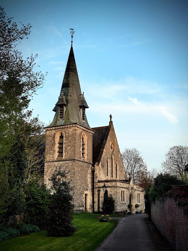 Stone church with a tall spire, surrounded by trees and a well-kept lawn under a clear blue sky.
