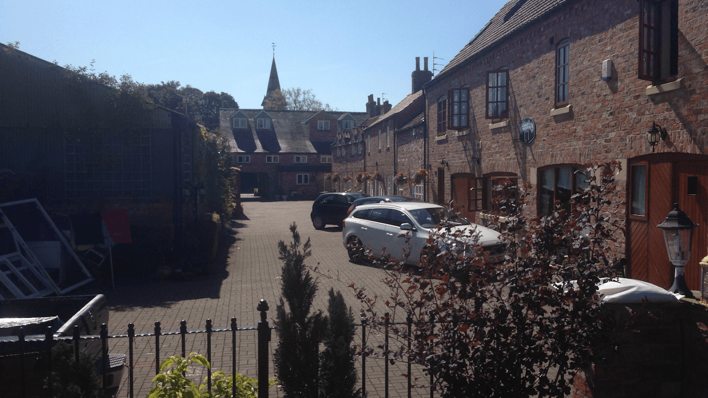 Quaint hotel courtyard with brick buildings, parked cars, and greenery under a clear blue sky.