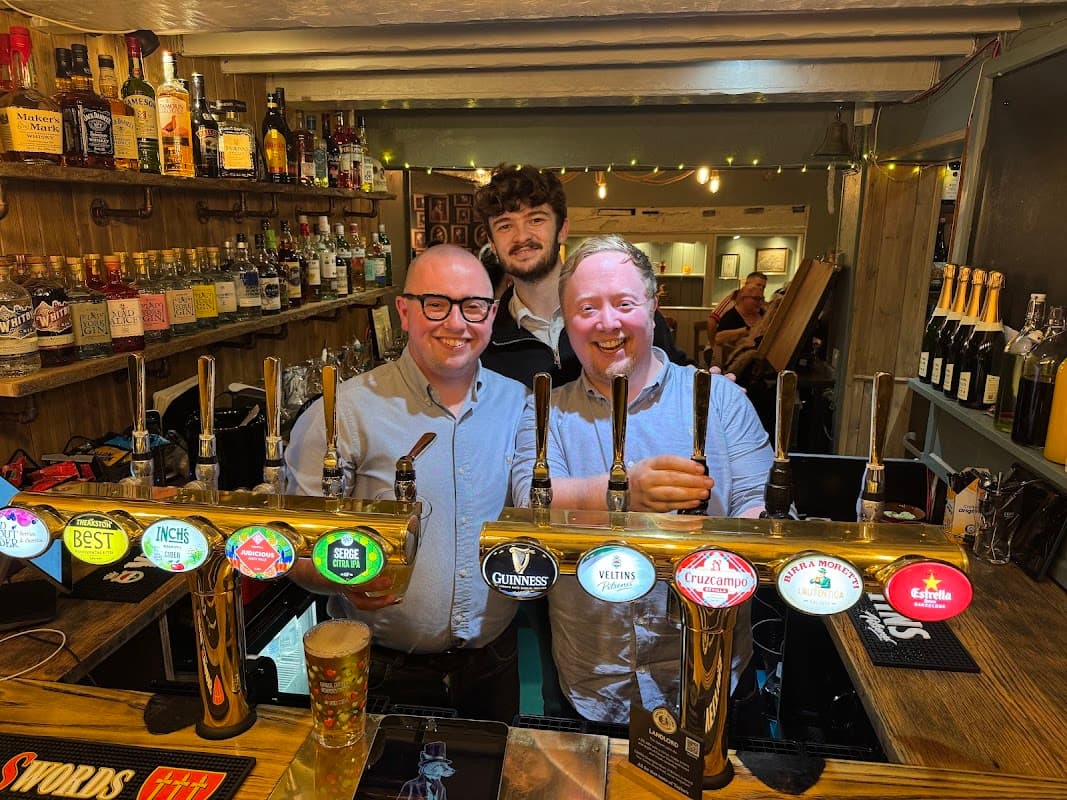 Two smiling men stand behind a bar with various beer taps and bottles, while a third man smiles in the background.